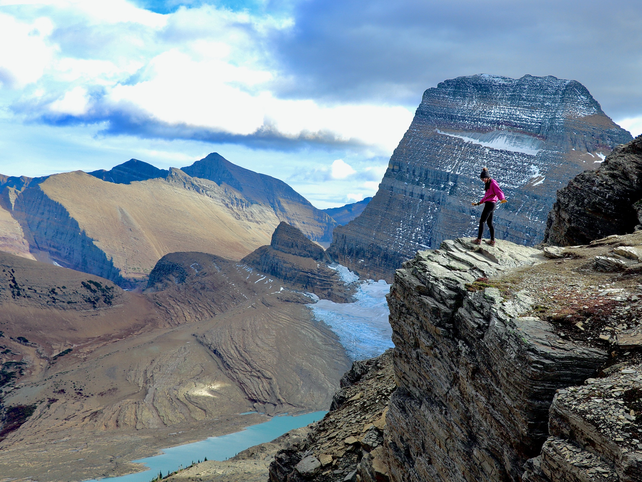 A hiker stands on a cliff edge above a glacial mountain valley